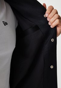 Black jacket interior showcasing a smooth fabric lining, metal snap closures, and a side pocket. A white t-shirt with a small logo is visible.