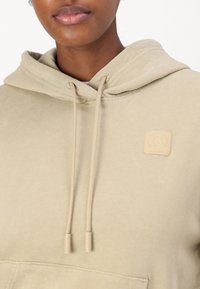 Beige huvtröja i mjukt tyg med en kenguruficka. Har en fyrkantig logotypapplikation och matchande dragskor.
