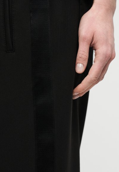 Μαύρο παντελόνι με λείο ύφασμα και γυαλιστερή πλαϊνή ρίγα. Η κοντινή λήψη δείχνει ένα χέρι που ακουμπά στη ραφή του ρούχου στο πλάι.