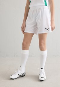 Calções atléticos brancos com cintura elástica, combinados com meias brancas até ao joelho e chuteiras de futebol pretas e brancas com riscas e logótipos.