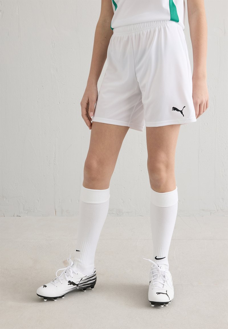 Calções atléticos brancos com cintura elástica, combinados com meias brancas até ao joelho e chuteiras de futebol pretas e brancas com riscas e logótipos.