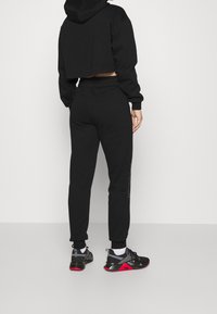 Svart kort hoodie och matchande joggers har ett mjukt, texturerat material. Joggers har sidfickor och diskret branding på benet. Svart och röda sneakers fulländar looken.