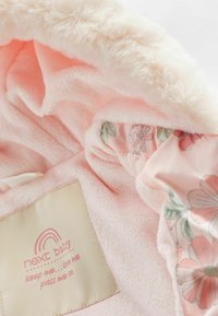 Blød rosa baby hoodie med fluffy futteret tekstur, blomsterfôr og et cremefarvet mærke med logo og plejeanvisninger.