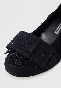 Μπλε σκούρο βελούδινο παπούτσι με χαμηλό τακούνι, διακοσμημένο με μεγάλο  φιόγκο με μαύρα καρφιά και στρογγυλεμένη μύτη. Ομαλή υφή με διακοσμητικά στοιχεία στο μπροστινό μέρος.