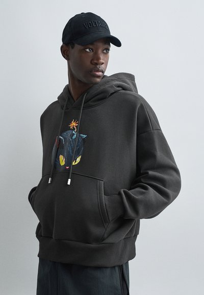 Hoodie noir avec une poche avant, graphique d'un chat en dessin animé avec des yeux jaunes éclatants et une explosion colorée, associé à une casquette noire.