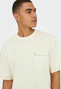 Cremefarvet t-shirt med kortærmer, kontrasterende sort syning, rund halsudskæring og en lille lomme på venstre bryst. Glat stofstruktur.