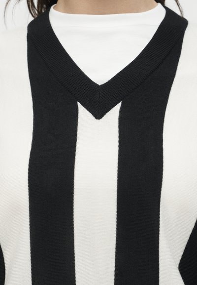 Λευκό πουκάμισο φορεμένο κάτω από μαύρο και άσπρο κάθετα ριγέ πουλόβερ με V-λαιμόκοψη, ορατό από τον λαιμό μέχρι το μέσο του στήθους.