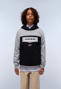 Sweat-shirt à capuche avec un dessus gris et un bas noir, présentant un écusson logo et un accent drapeau. Porté par-dessus une chemise à col rayée bleue.