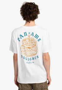 Bílý bavlněný tričko s barevným grafickým designem na zadní straně, zobrazující naskládané dezerty s textem "PANAME DELIGHTS" v modré a zlaté barvě.
