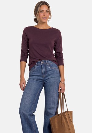 Femme aux cheveux châtain clair attachés porte une chemise violette à manches longues et un jean bleu, tenant un grand sac cabas en cuir marron.