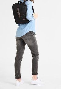 Black fabric backpack with a rolled top, brown strap closure, worn over a light blue shirt, paired with grey jeans and white sneakers.