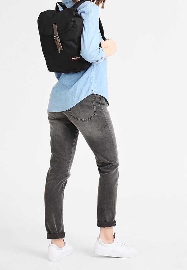 Black fabric backpack with a rolled top, brown strap closure, worn over a light blue shirt, paired with grey jeans and white sneakers.