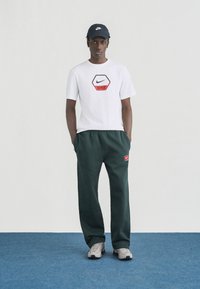 Άνδρας που φοράει λευκό t-shirt Nike, σκούρο πράσινο παντελόνι φόρμας Nike, γκρι αθλητικά παπούτσια και μαύρο καπέλο Nike, στέκεται σε μπλε χαλί μπροστά σε λευκό τοίχο.