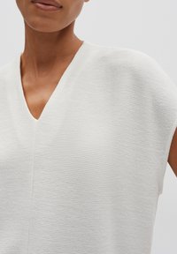 Haut blanc texturé avec un col en V, des manches courtes et de subtiles rayures horizontales, mettant en valeur un tissu léger et respirant.