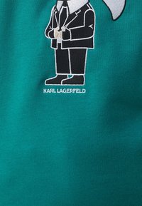 Sudadera color teal con una figura de cartoon bordada en traje negro y camisa blanca, sosteniendo unas gafas. El texto debajo dice "KARL LAGERFELD."