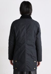 Osoba s krátkými tmavými vlasy, oblečená v dlouhém černém kabátu s mosaznými knoflíky na zadní straně, stojící proti jednoduchému světlému pozadí.