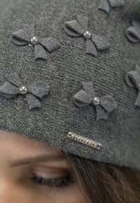 Cappello di lana grigio adornato con fiocchi in feltro e perle. Presenta un'etichetta logo metallica sul lato. Texture morbida, vestibilità aderente.