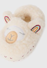 Chausson en peluche beige en forme d'ours, avec des yeux et un nez brodés, des accents d'oreilles colorés et une surface texturée avec de petits motifs rouges.