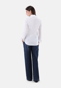 Weiße Bluse mit Kragen und langen Ärmeln, kombiniert mit hochgeschnittten, weiten blauen Jeans, glatte Textur und klassischer Schnitt.