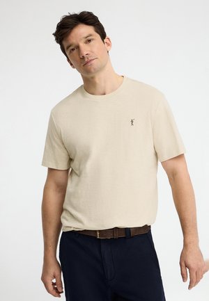 Homme aux cheveux foncés portant un t-shirt beige avec une petite figure brodée et un pantalon bleu marine avec une ceinture marron, debout devant un fond uni.