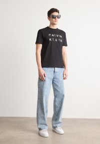 Tricou negru din bumbac cu logo-ul "CALVIN KLEIN" în alb, asortat cu blugi denimi deschiși, cu picioare largi și adidași albi. Modelul poartă ochelari de soare.