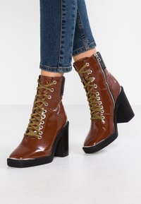 Bottines en cuir verni marron avec un talon épais noir, des lacets verts et une fermeture éclair latérale. Caractérisées par un bout rond et des accents contrastants.
