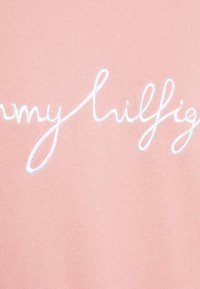 Tissu rose avec texte en cursive brodé en blanc indiquant « Tommy Hilfiger » sur une section en gros plan d'un vêtement.