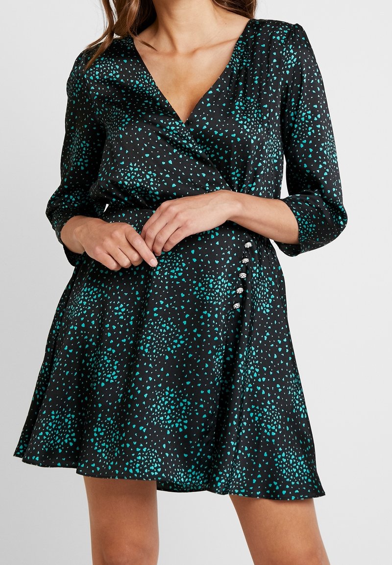 Robe portefeuille noire avec un motif de points dispersés teal, manches trois-quarts, et boutons décoratifs argentés sur le côté de la taille.