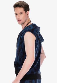 Ärmellose blaue Kapuzenjacke mit Camouflage-Muster, Kordelzug am Hals und kontrastierendem schwarzen Saum. Hergestellt aus einem weichen Stoff.