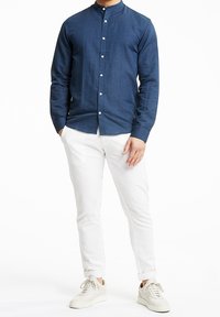 Marineblauwe linnen overhemd met lange mouwen en kraag, voorzien van een knopenlijst, gecombineerd met witte slim-fit broek en beige sneakers.