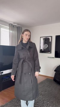 Trench coat cinzado cinza escuro com grandes botões, golas largas e corte reto, usado sobre uma blusa preta e jeans claros. Textura suave.
