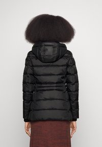 Doudoune noire avec capuche, présentant un design matelassé, des poignets élastiques et une silhouette ajustée. Tissu texturé avec des coutures visibles.