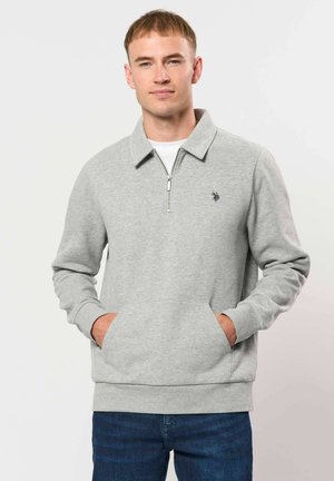 Jeune homme portant un sweat-shirt zippé gris clair avec col et poches avant, associé à un jean bleu, debout devant un fond uni.