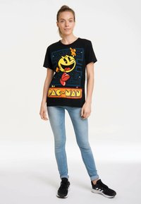 Black short-sleeve T-shirt featuring a colorful Pac-Man graphic, with blue maze background and bold "PAC-MAN" lettering.