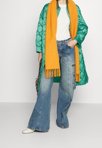 Cappotto lungo trapuntato verde, t-shirt bianca, sciarpa di lana arancione, jeans blu a gamba larga e scarpe color crema. Una borsa a tracolla marrone completa l'outfit.