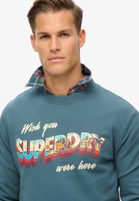 Sudadera azul de cuello redondo, con un gran texto gráfico colorido de 'SUPERDRY' y un cuello de camisa de cuadros superpuesto.