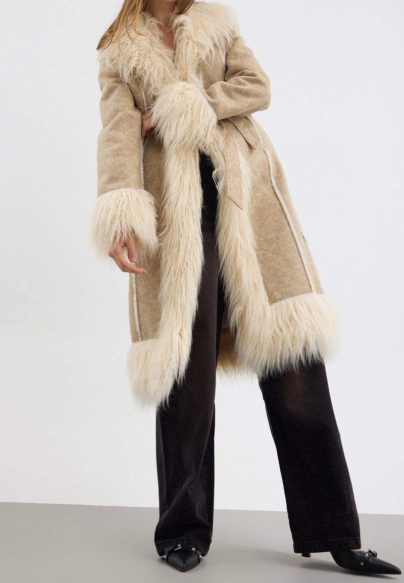 Femme portant un long manteau en daim beige avec une bordure en fourrure crème, un pantalon noir à jambes larges et des chaussures noires pointues, se tenant devant un fond blanc.