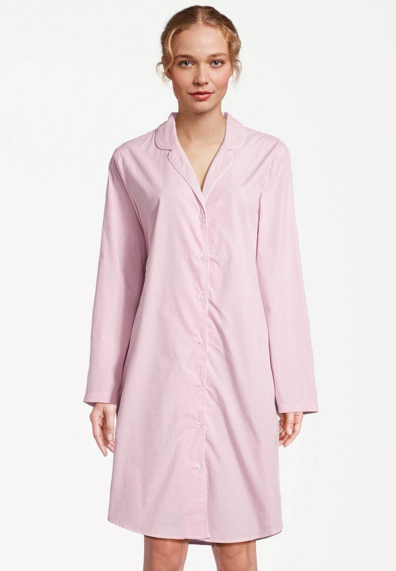 Femme portant une chemise de nuit rose clair, à manches longues, boutonnée jusqu'aux genoux avec un col, debout devant un fond blanc uni.