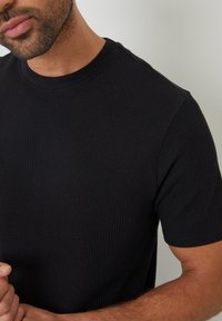 Schwarzes kurzärmliges T-Shirt aus strukturiertem, geripptem Stoff. Es hat einen klassischen Rundhalsausschnitt und ein figurbetontes Design.