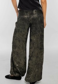 Pantalon large vert métallisé à surface texturée avec une ceinture élastique, associé à des ballerines noires ornées de clous.