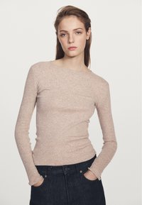 Dlouhý rukáv, přiléhavý ribbed top v měkké béžové látce, s širokým výstřihem, spárovaný s tmavými džíny, s rukama v kapsách.