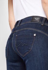 Mörkblå denimjeans med en figurnära design, som har distinkta bakfickor med rundad söm och en svart varumärkesetikett vid midjan.