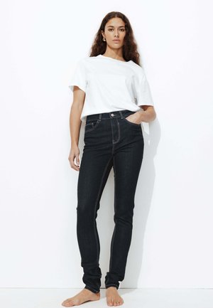 Jean skinny en denim sombre avec des coutures contrastantes, taille haute et agencement standard à cinq poches, associés à un t-shirt blanc basique.