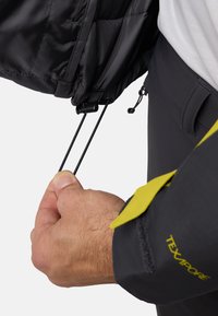 Giacca isolante nera con cordino regolabile. Caratterizzata da un polsino grigio con dettagli in logo giallo. Cordino di regolazione manuale visibile.