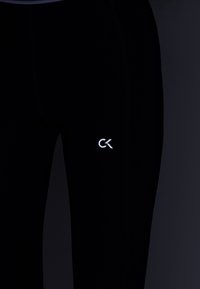 Close-up van zwarte strakke leggings met een klein wit "CK" logo op de bovenbenen tegen een donkere achtergrond.