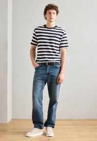Gestreiftes kurzärmliges T-Shirt in Marineblau und Creme, kombiniert mit hellblauen Jeans und cremefarbenen Sneakers. Ein schwarzer Gürtel hebt das Outfit hervor.