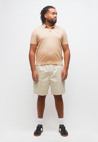 Άντρας με ντραντλοκς, φορώντας μπεζ κοντομάνικη πόλο μπλούζα, ανοιχτό μπεζ σορτς, λευκές κάλτσες και μαύρα αθλητικά παπούτσια, στέκεται μπροστά σε λευκό φόντο.