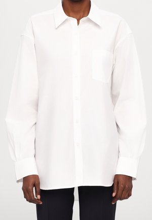 White long-sleeve button-up shirt with a pocket on the left chest worn over black pants, shown from neck to hips.