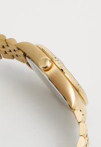 Guldfarvet metalarmbåndsur med flad kasse, rund urskive, tekstureret bezel og tekstureret guldkæde. Kronen er placeret på siden.