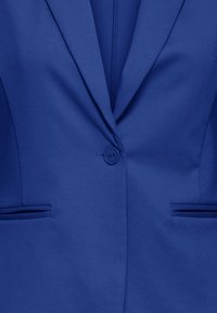 Suur plaan sinisest bleiserist, mille esiküljel on kinnitatud üks nupp ja kaks horisontaalset lõhikutaskut.